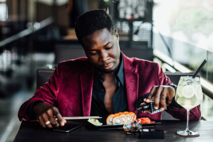 Masturdating: Is it self-care? - cheerful curly african guy holding chopsticks sushi rolls chinese food fish restaurant terrace - Shujaa Designs